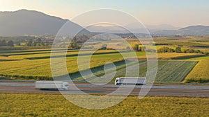 AERIAL: Flying along a white semi truck hauling freight down a scenic highway.