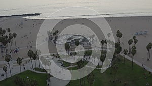 Aerial: flying away from Venice Beach Skatepark with Visitors and Palm Trees, Sunset in Los Angeles