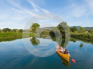 AERIAL: Flying behind tourists canoeing along calm river running past a church