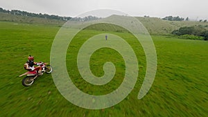 Aerial follow view two enduro motorbike riders uphill driving on green grass hill field