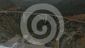 Aerial footage of Bixby Creek Bridge spanning valley at sea coast. Waves rolling on sand. Hazy view of mountain