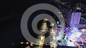 aerial footage of the Carolina Beach Boardwalk with a Ferris wheel, colorful amusement rides, people walking, a sandy beach