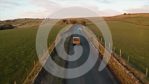 Aerial footage following a vehicle with a dog in the back. Narrow farmland road