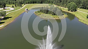 Aerial footage of a fountain in a pond in Whitecourt rotatory park with green grass and fir trees