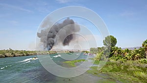 Aerial footage of the green waters of the Colorado River with people in boats and smoke from a brush fire on the banks of the rive