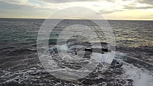 Aerial footage of the vast blue ocean water with waves crashing into the rocks with powerful clouds at sunset at Victoria Beach