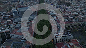 Aerial footage of wide boulevard with trees in town development. La Rambla street and Placa Reial at dusk. Barcelona