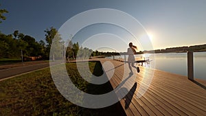 Aerial FPV drone view of sporty young woman in activewear running along wooden pier around lake on bright sunny summer