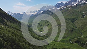 AERIAL: Green landscape with sunlit forested hills and high snowy mountains