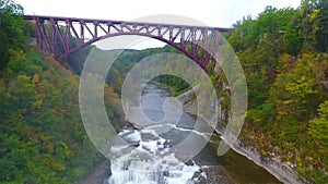 Aerial high to low by steel maroon bridge over stunning raging waterfall cutting through layered cliffs in lush green