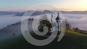 Aerial landscape of mountain hills coverd in fog and Saint Tomas church, Slovenia on sunset