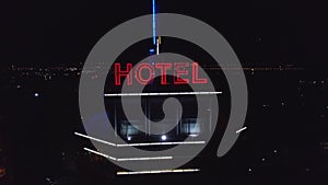 Aerial: neon red inscription hotel on the roof of a modern building in the night city. Traffic cars are next on the road