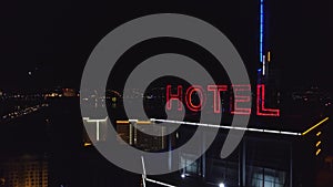 AERIAL: Neon red inscription hotel on the roof of a modern building in the night city. Traffic cars are next on the road
