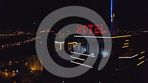 AERIAL: Neon red inscription hotel on the roof of a modern building in the night city. Traffic cars are next on the road