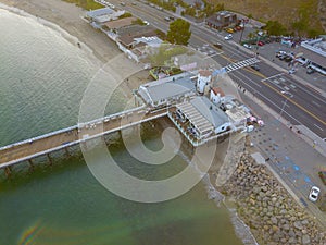 An aerial overhead shot of a pier over the green ocean water surrounded by lush green trees, parked cars on a street