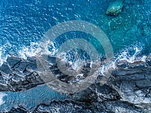 Aerial overhead top view of ocean mediterranean sea waves reaching and crashing on rocky shore beach, near travel