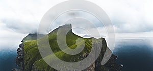 Aerial panorama of a small white lighthouse located on the edge of a huge cliff and the island of Kalsoy. Kalsoy is an