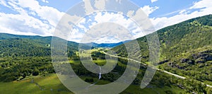 Panoramic banner view of landscape with mountains, green trees, field, road and river under blue sky and clouds in summer