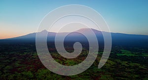 Aerial, panoramic view on Mount Kilimanjaro volcano, summit covered in snow lit by first sun rays with masai villages with