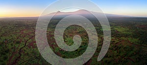 Aerial, panoramic view on Mount Kilimanjaro volcano, summit covered in snow lit by first sun rays with masai villages with
