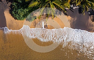 Aerial photo of surfer lying next to surfboard on the sandy ocean shore in STAR pose and sun tanning under tall palm trees. Soft