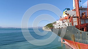 Aerial for red, empty barge moored along the sea shore near a slope covered with green trees on blue sky background