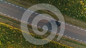 Aerial shot from above driving black car in a field along a rural road in summer at sunset.