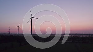 Aerial shot by a drone of windmills with rotating wings among green fields against a sunrise or sunset background. Wind
