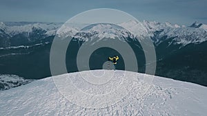 Aerial shot flying over winner male snowboarder admiring mountain landscape from peak of rock