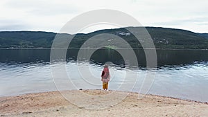 Aerial shot. Happy tourist woman with a big backpack stands overlooking the lake and looks at nature. Back view.