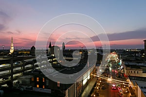 Aerial shot of office buildings and tower cranes in the city skyline with lights and cars driving along the street at sunset