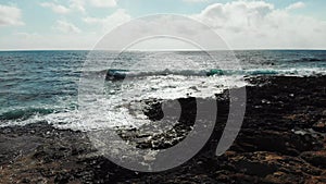 Aerial shot over sea waves hitting rocky beach splashing against rocks. Blue sky with sun road on the horizon. Ocean on a windy da