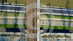 Aerial shot of passenger train moving on railway bridge across the river and bicycle road, top view. Ecologic