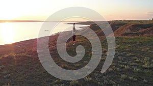 Aerial shot of young sporty woman making exercises on the egde of slope near the lake or liman during beautiful sunrise