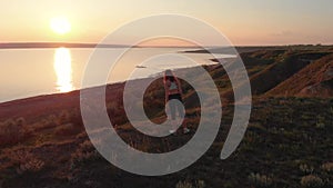 Aerial shot of young sporty woman making exercises on the egde of slope near the lake or liman during beautiful sunrise