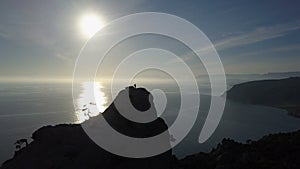 Aerial silhouette of young woman on the top of a mountain facing the sea. Flight over Lady on the summit raising up