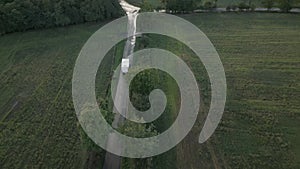 Aerial slow movement around car driving on road surrounded by greenery fields and dense trees