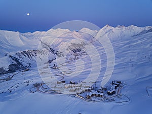 Aerial of snowy mountain range on winter sunrise at ski resort. Moon above mountains valley and village with switchbacks