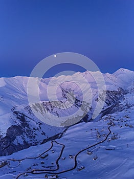 Aerial of snowy mountain range on winter sunrise at ski resort. Moon above mountains valley and village with switchbacks