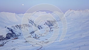 Aerial of snowy mountain range on winter sunrise at ski resort. Moon above mountains valley and village with switchbacks