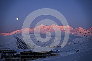 Aerial of snowy mountain range on winter sunrise at ski resort. Moon above mountains valley and village with switchbacks