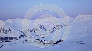 Aerial of snowy mountain range on winter sunrise at ski resort. Moon above mountains valley and village with switchbacks