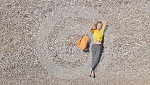 Aerial top view happy woman traveler in yellow shirt lying alone on Empty pebble beach and waving hand to camera.