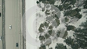 Aerial top view of a highway in winter weather with driving transport. Video. Snow covered ground and pine tree forest