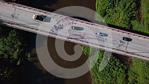 Aerial top view of a narrow bridge above brown dirty river. Clip. Green shores with bushes and driving cars.