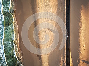 Aerial vertical view of yellow tropical sandy beach with black long way road and car traveling - ocean blue waves and shore -