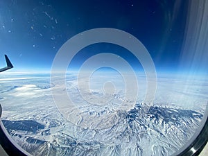 An aerial view from an airplane window of mountains, snow, clouds and blue skies