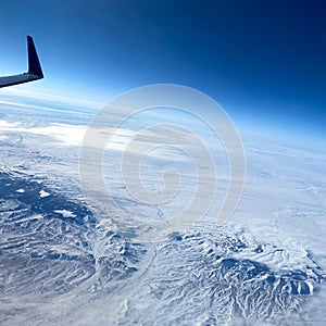 An aerial view from an airplane window of mountains, snow, clouds and blue skies