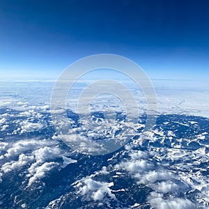 An aerial view from an airplane window of mountains, snow, clouds and blue skies