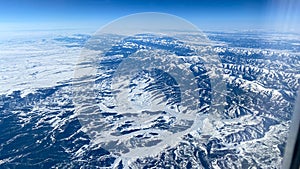 An aerial view from an airplane window of mountains, snow, clouds and blue skies